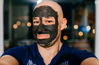 Yüzünde kömür maskesi olan bir adam karanlık arka planda ışıklarla selfie çekiyor..