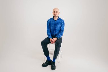 Mavi gömlekli, siyah pantolonlu ve mavi çoraplı yakışıklı beyaz arka planda izole edilmiş uzun bir sandalyede oturuyor..