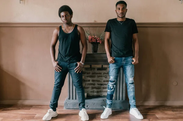 Two handsome adult african male friends in casual clothing posing together like gangsters in front of decorative fireplace on wall. Two confident african american men indoor lifestyle portrait
