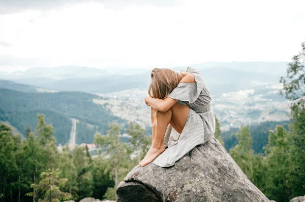 Young girl sits at stone on top of mountain and hugs her legs with head lying on knees. Beautiful blonde woman in depression hiding her face. Psychological female outdoor portrait. Loneliness concept
