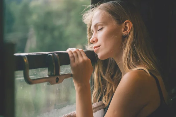 Mood atmospheric lifestyle portrait of young beautiful blonde hair girl looking out of window from riding train. Pretty teen enjoying beauty of nature from moving train car in summer. Travel concept.