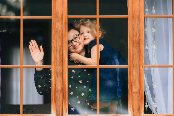 Portrait of beautiful young caucasian female with pretty face, short dark hair, big eyes, glasses with her cheerful child and looks through big window