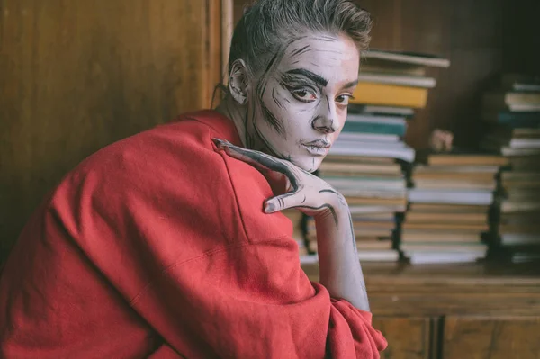 Stylish young  girl with cartoon body art sitting against vintage wooden cupboard with books.