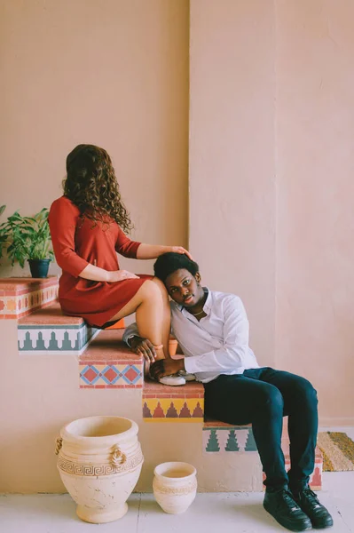 Weird interracial couple. Happy adult smiling dark skinned man siiting on stairs in decorative room and hugging legs of his caucasian girlfrined with face covered by hair. Portrait of strange lovers