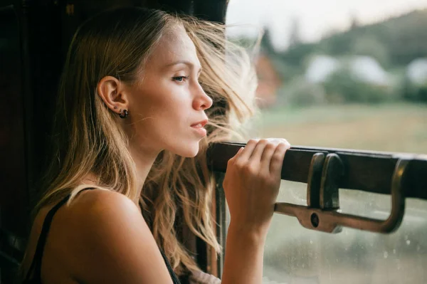 Mood atmospheric lifestyle portrait of young beautiful blonde hair girl looking out of window from riding train. Pretty teen enjoying beauty of nature from moving train car in summer. Travel concept.