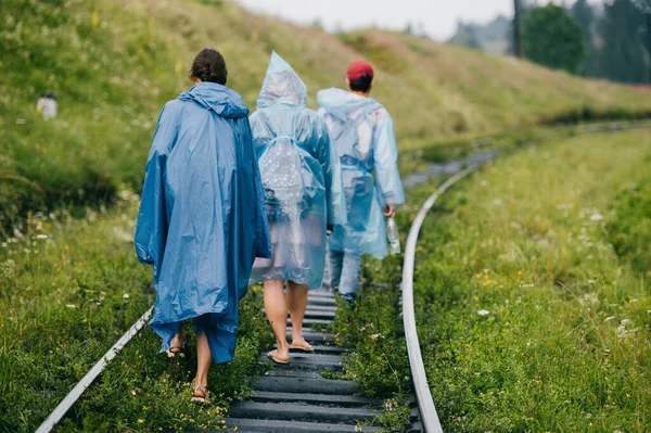 Crew of informal young travelers in raincoats walking in danger zone on railroad outdoor at nature. Company of friends rest on vacation. People go on railway sleepers. Cloudy muggy weather