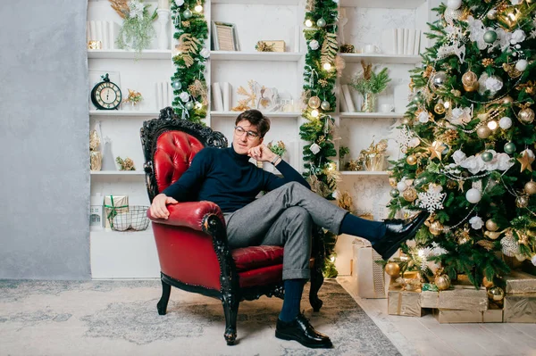 スタイリッシュなエレガントな男は 新しい年の装飾とスタジオでクリスマスツリーの近くの椅子に座って — ストック写真