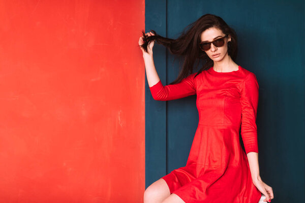 Beautiful fashion girl with long hair, spanish appearance in sunglasses and red elegant dress posing on blue red wall in studio. Amazing babe in suglasses pulling her black hair. Hairstyle advertsing