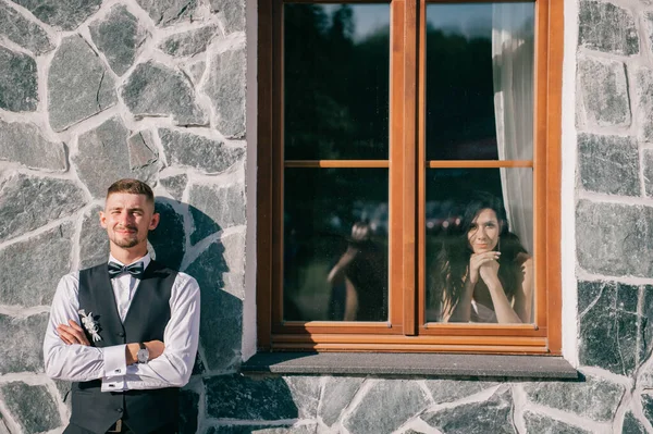 エレガントな新郎が石の壁の前に立っている彼の花嫁は窓から彼を見て — ストック写真