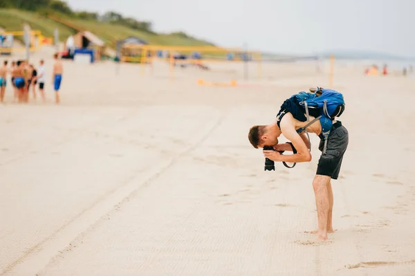 一个穿着短裤 背着背包 赤身裸体的年轻人 在拍摄沙子的时候靠在后面调整相机的镜头 摄影记者寻找好的相框 — 图库照片