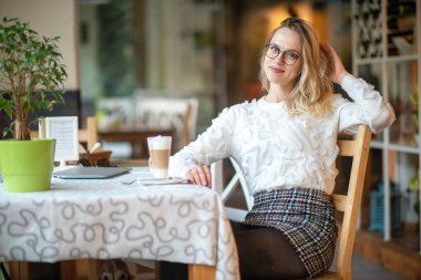 Beyaz süveterli, gözlüklü, kafede oturan ve kameraya pozitif yüz ifadesiyle poz veren güzel sarışın kadın..