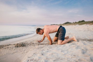 Genç, çıplak ayaklı, şortlu, dizlerinin üzerinde duran ve yaz akşamları denizin ötesindeki kumsalda el pençe divan kazan adam. Okyanus çocuğu gibi ıssız bir yerde kumla sahilde oynayan komik çocuk.. 