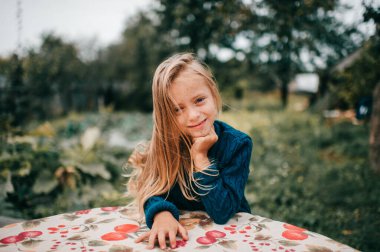 Mavi kapüşonlu, uzun sarı saçlı, güzel beyaz kız ağacın yanındaki çarşafta uzanıyor, gülümsüyor ve saçlarıyla oynuyor.