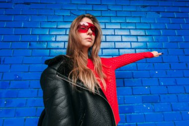 Uzun saçlı, siyah koyun derisi ceketli, açık havada mavi tuğla duvarın üzerinde duran kırmızı gözlüklü, esmer bir kadının moda portresi..