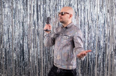 Saçsız, siyah güneş gözlüklü, parlak gömlekli, siyah deri pantolonlu ve gri spor ayakkabılı güzel beyaz adam küçük gümüş bir mikrofonda şarkı söylüyor..