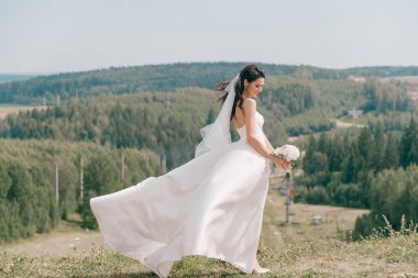 Beyaz gelinlikli güzel gelin doğada bir tepede poz veriyor.