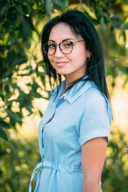 Büyük kahverengi gözlü, uzun siyah saçlı, kısa mavi elbiseli, siyah gözlüklü sevimli beyaz kız gülümseyip bahçedeki yeşil ağacın yanında seviniyor.