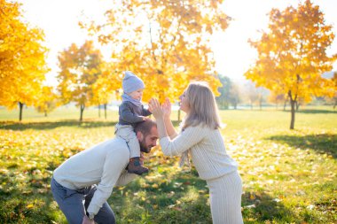 Mutlu anne, sonbahar parkında babasının omuzlarında bebeğiyle el çırpıyor.