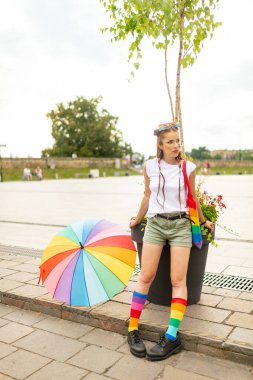 Yüzünde lgbt gökkuşağı olan şık biseksüel kız şehir meydanında renkli şemsiye ve çantayla poz veriyor..