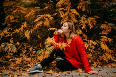 Güzel beyaz kadın, kırmızı kazaklı, siyah pantolonlu ve renkli örgü şapkalı, ağacın altındaki sarı çimlerin üzerinde oturuyor, gülümsüyor ve eğleniyor.