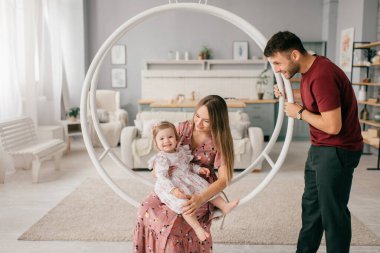 Genç, beyaz bir anne ve güçlü bir baba parlak bir odada küçük tatlı kızıyla birlikte yuvarlak bir salıncakta oturup eğleniyor.