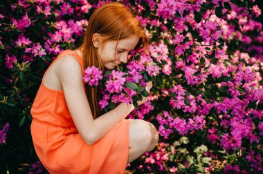 Uzun kızıl saçlı, soluk tenli, etkileyici genç kız bahçede çiçekleri kokluyor..