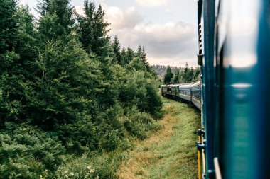Yazın trenle gezmekten pencere dışındaki manzara. Tepeler, dağlar ve ormanlar. Tatil ve seyahat konsepti. Tren vagonlu lokomotif tren yolu boyunca ilerliyor.