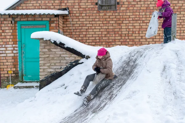 一个快乐的女孩用塑料袋当雪橇 骑着车从地窖里下来 带着冰凉的远足 来到砖村的房子前 — 图库照片