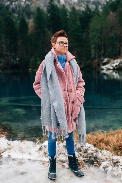 Picture of handsome woman in warn pink coat, denim trousers and with long grey scarf stands near the beautiful lake and smiles
