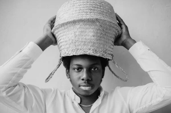 Closeup lifestyle portrait of strange young african black man holding straw basket above his head on textured wall background and looking at camera. Dark-skinned boy in white t-shirt funny portrait.