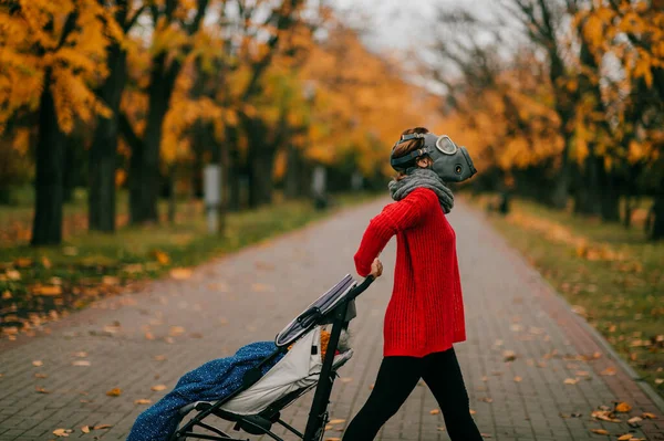 A beautiful tall woman in a red jacket and black pants with a gas mask on her head went for a walk with a child in a pram.