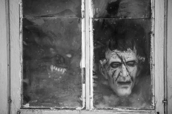 Black and white picture of a child in a terrible monster mask peeks out of an old dirty window in winter