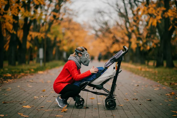 在秋天的公园里 一个穿着红色夹克 头戴防毒面具 穿着黑色裤子的漂亮高个子女人带着一个孩子在一个婴儿车里散步 — 图库照片
