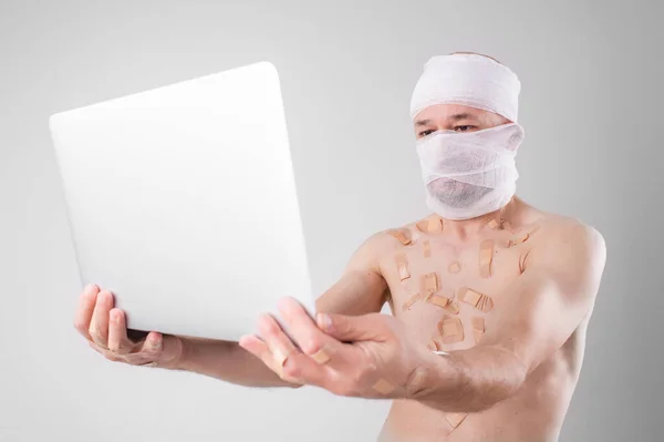 Injured man with many patches on his body and bandaged head serfing internet with laptop isolated on white background