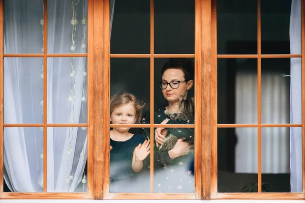 Portrait of beautiful young caucasian female with pretty face, short dark hair, big eyes, glasses with her cheerful child and looks through big window