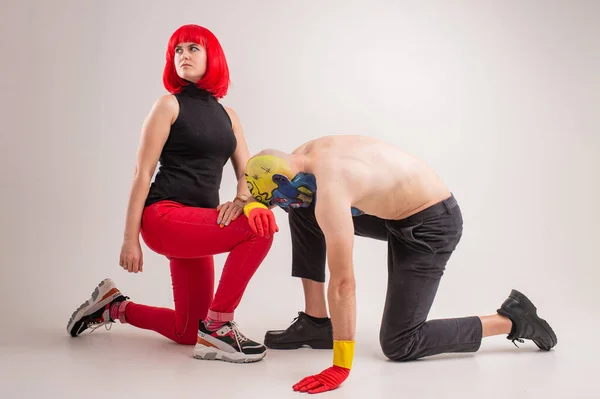 A strange man with makeup on his head and shoulder in red-yellow gloves, black pants and boots, no T-shirt sits on a floor with young caucasian female with red hair, red trousers, black t-shirt