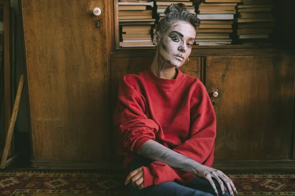 Stylish young  girl with cartoon body art sitting against vintage wooden cupboard with books.