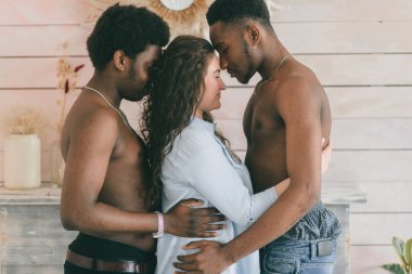 Çok kültürlü aşk ve ilişkiler kavramı. Beyaz seksi kız iki üstsüz koyu tenli adamı kucaklıyor. Irklar arası aşk üçlüsünün kapalı bir portresi. İki tutkulu Afrikalı adam sevgililerine sarılıyor.