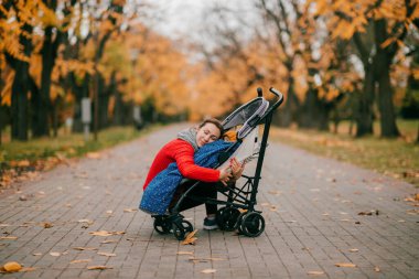 Kırmızı ceketli ve siyah pantolonlu yakışıklı beyaz kadın sonbahar parkında bir bebekle yürüyüşe çıkıyor..