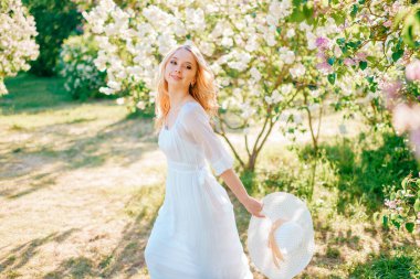 Beyaz leylak ağacının yanında uzun mavi elbiseli genç beyaz kadın duruyor.