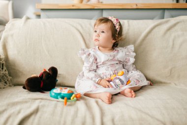 Çekici elbiseli küçük beyaz kız kanepeye oturur, çeşitli oyuncaklarıyla oynar ve gülümser.