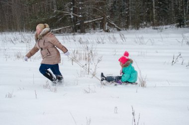 Anne kızıyla birlikte kızağını çekiyor. Kışın doğada karlı bir günde üzerinde oturuyor..