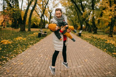 Beyaz ceketli, siyah pantolonlu ve gri ayakkabılı güzel beyaz anne, sonbahar parkında oğluyla oynuyor..