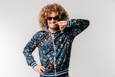 80 'lerin moda eşofmanlı komik bir adamın kapalı portresi çiçek desenli ve güneş gözlüklü parmaklarıyla dişlere dokunan. Yetişkin, neşeli, kıvırcık saçlı, beyaz stüdyo geçmişiyle dalga geçen bir erkek.