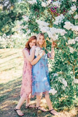 Leylak bahçesinde sarışın ve kahverengi saçlı iki güzel kız birlikte vakit geçirip gülümsüyor.