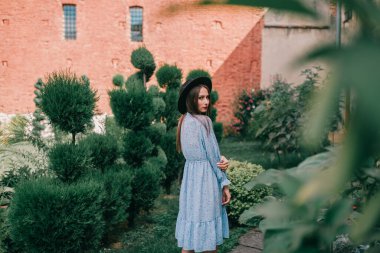 İkili mavi elbiseli, uzun saçlı esmer kız yeşil bahçede Fransız tarzı poz veriyor.