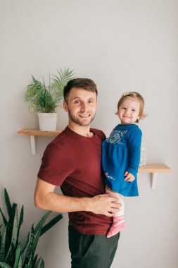 Beyaz tenli yakışıklı, güçlü bir baba, parlak bir odada, rafta oturan küçük tatlı kızını tutuyor.
