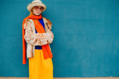 Kırmızı gözlüklü sıra dışı bir genç mavi süveter, sarı etek ve uzun turuncu atkı ve beyaz şapkalı bir ceketle yürüyüşe çıktı. Mavi arka plandaki kamera için farklı pozlar gösteriliyor..