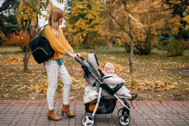Büyük mavi gözlü, kısa kızıl saçlı, güzel mutlu annesiyle sonbaharda bebek arabasında dudaklarını pompalayan güzel bir bebek.