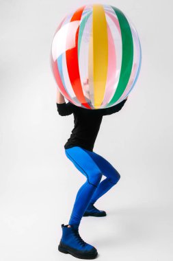 Spor kıyafetleri içinde şık bir adam, plaj topunu suratının önünde beyaz stüdyo arka planında tutuyor..